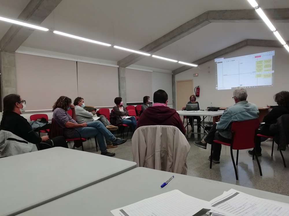 Aula formativa amb persones assegudes en cercle i la formadora davant el projector